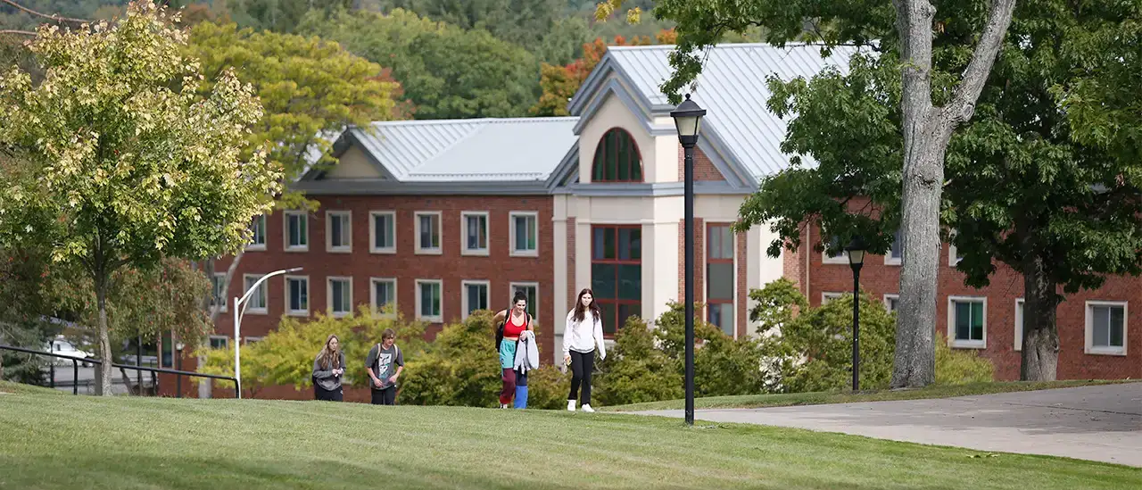 People walking on a sidewalk amongst green grass and lush trees with a residence hall is in the background
