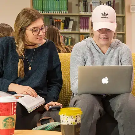 Two students sitting together and working on a project or studying, one with a book and the other with a laptop