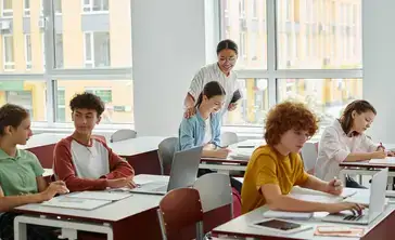 Smiling teacher standing next to adolescent students during lesson with devices in class