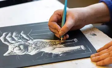 Student works with a calligraphy stylus to draw a white cat on a black board.