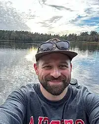 Dr. Alex Sotola smiling with a lake shoreline and sunlit cloudy sky behind him.