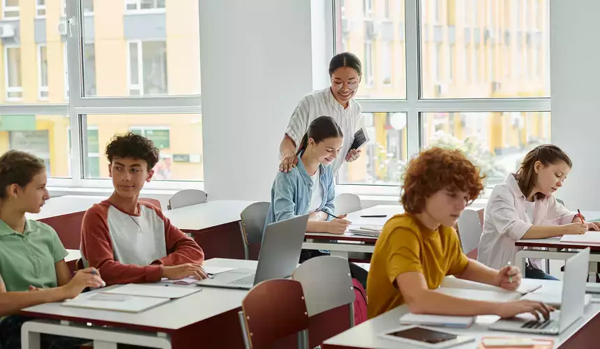 Smiling teacher standing next to adolescent students during lesson with devices in class