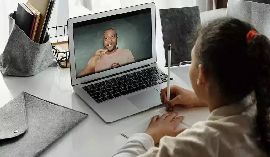 A child takes notes while watching their teacher on a laptop