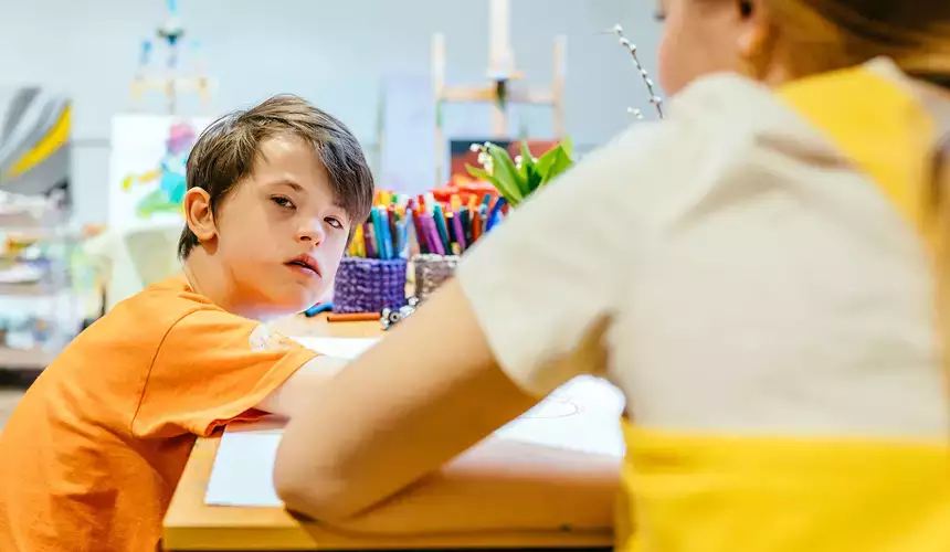 Rear view of teacher sitting with boy with special needs looking at camera drawing