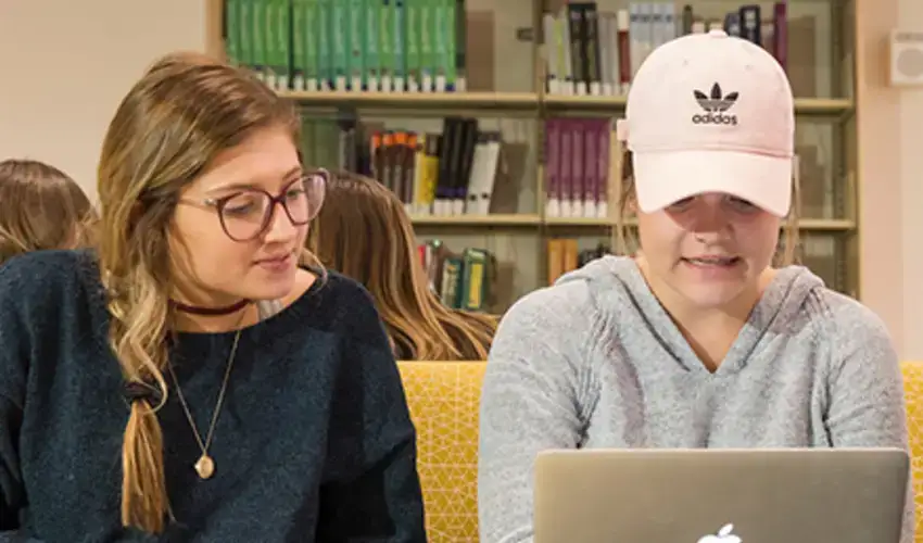 Two students sitting together and working on a project or studying, one with a book and the other with a laptop
