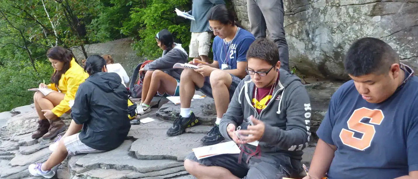 Students take notes and make sketches while seated on a rock outcropping.