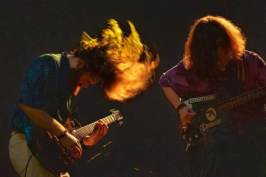 Guitarist of band Buffalo Joe and the Hobos plays and tosses his hair, backlit by stage lights.