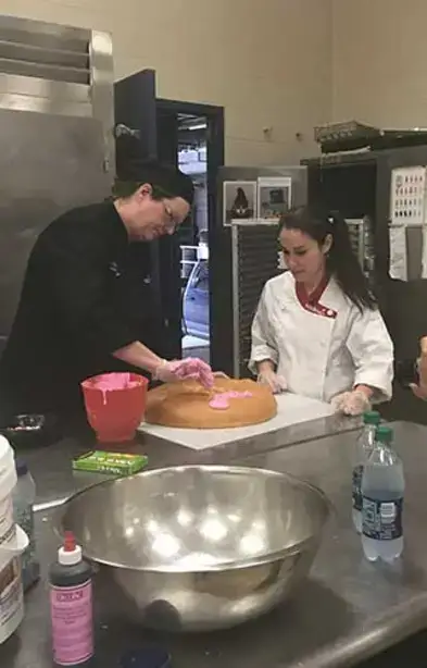 Julia and a helper apply pink frosting to a giant donut in the kitchen.