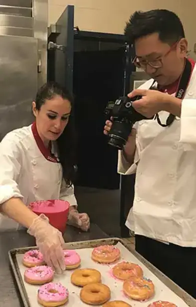 Julia frosts donuts with JP taking photos in the kitchen at Wilsbach Dining Hall.