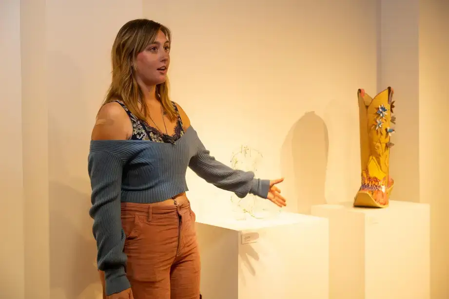An art gallery student exhibitor standing near their cowgirl boot sculptures (one appears to be made of a wire form and the other decorated cardboard)