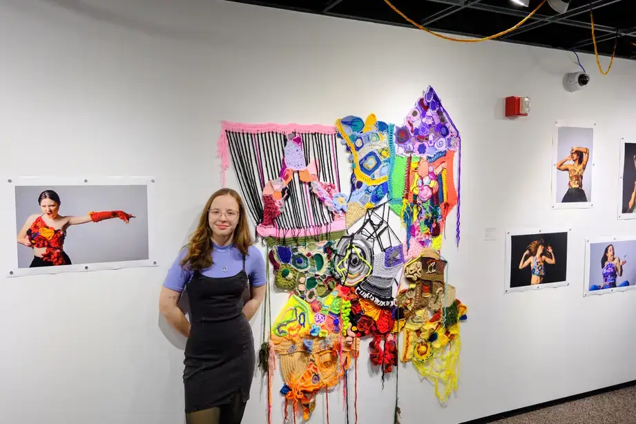 An art gallery student exhibitor standing near their colorful display containing a collage of yarn art and photos.