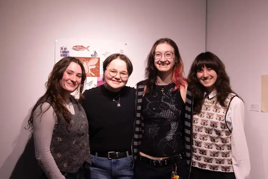 A group of art gallery student exhibitors posing together for a photo in the art gallery (a portion of an exhibit is seen behind them)
