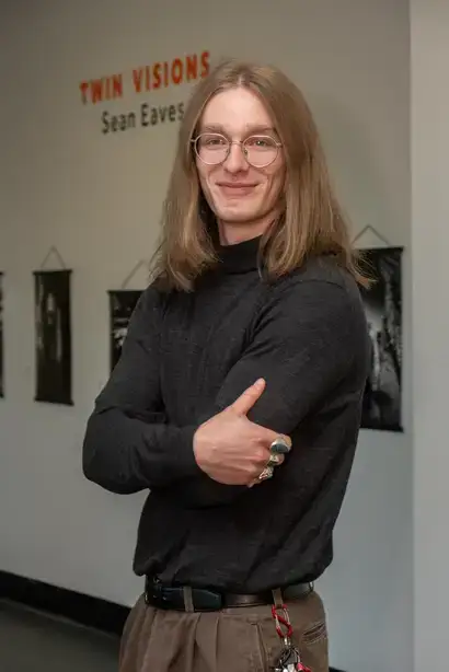 Sean Eaves, an art gallery student exhibitor standing in front of some of their pieces with "Two Visions, Sean Eaves" written on the wall behind them.