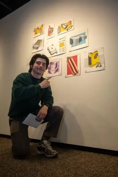 An art gallery student exhibitor kneeling and pointing to their a collage of pieces in their exhibit containing fine art drawings