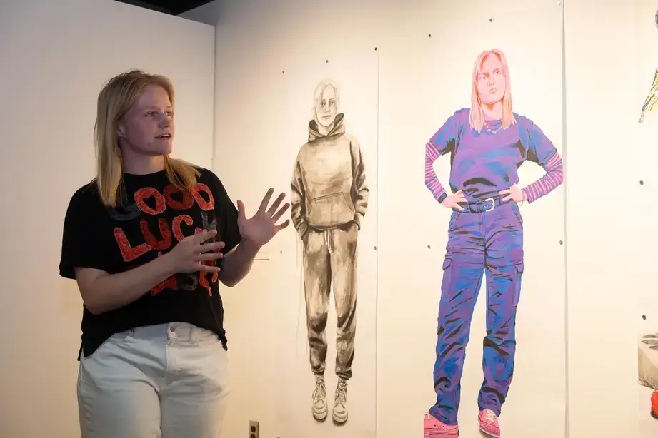 An art gallery student exhibitor standing near their exhibit containing life size fine art drawings of themselves (one in black and white, one in color).