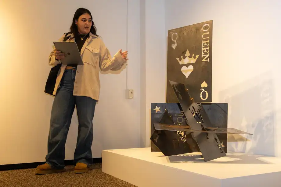 An art gallery student exhibitor standing near their exhibit containing metal sculptures that appear to be playing cards (Queen, Joker, King, Jack, etc.)