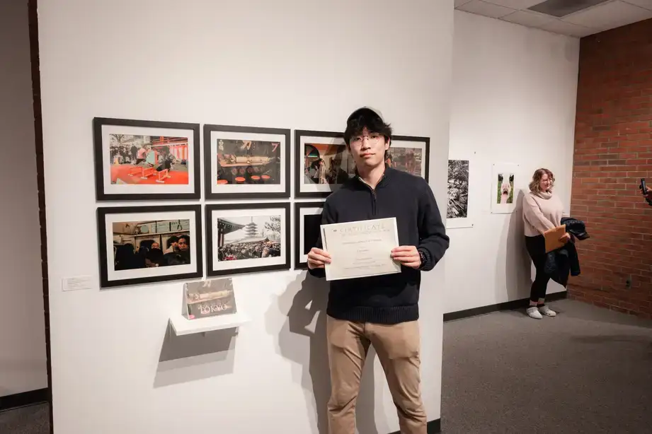 An artist posing with the Certificate of Achievement in front of their exhibit during a Juried Student Show