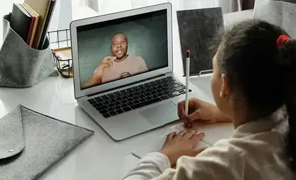 A child takes notes while watching their teacher on a laptop