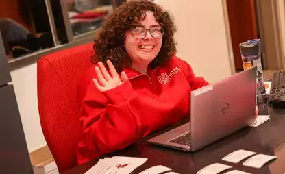 Person sitting at a desk smiling and waving "hi"