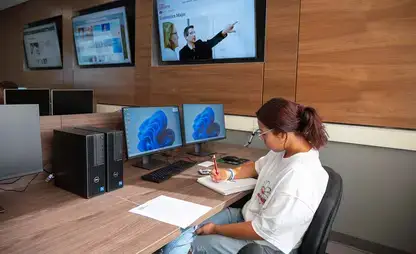 Student takes notes while seated at computer station in a classroom.