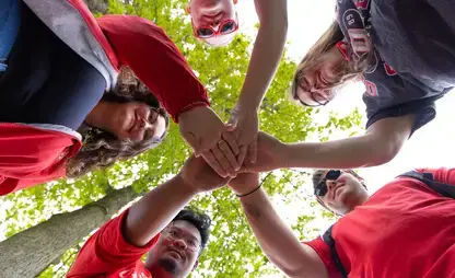 photo shot from the ground looking up through 5 students standing in a circle with their hands meeting in the middle