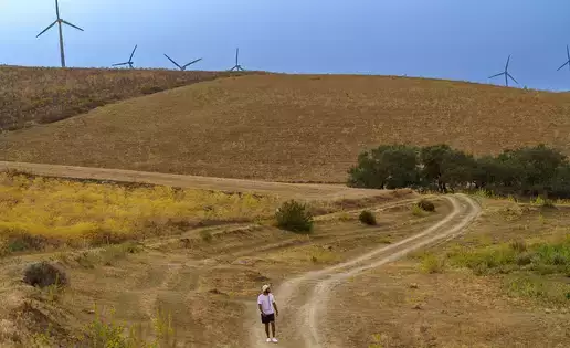 An individual walks along a dirt road with wind turbines seen off in the distance