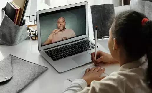 A child takes notes while watching their teacher on a laptop