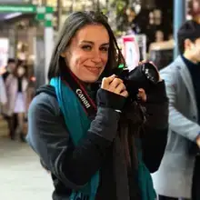 Julia Goolia holds a camera and smiles posing on a busy street.