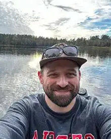 Dr. Alex Sotola smiling with a lake shoreline and sunlit cloudy sky behind him.